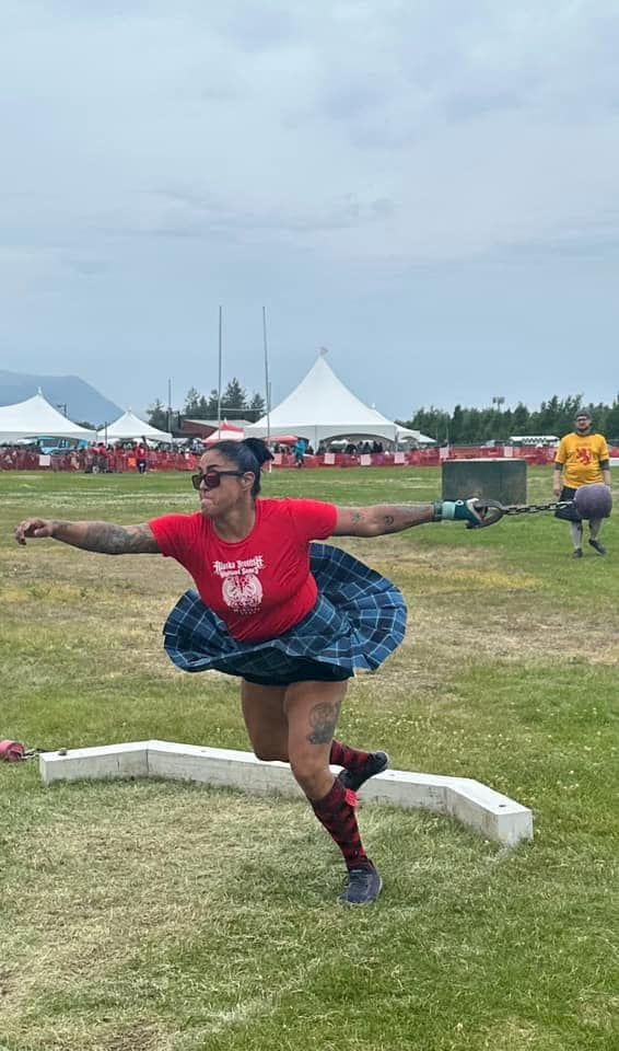 Woman throwing a caber at an outdoor Highland games competition. She's wearing a red shirt, kilt, and sunglasses.