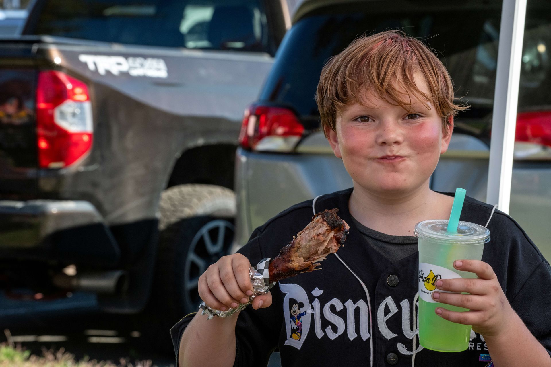 Smiling child holding a turkey leg and a green drink outdoors near parked cars