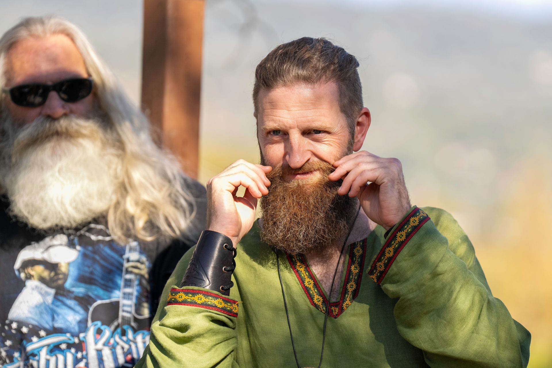 Two bearded men outdoors, one smiling and holding his mustache, both in colorful clothing