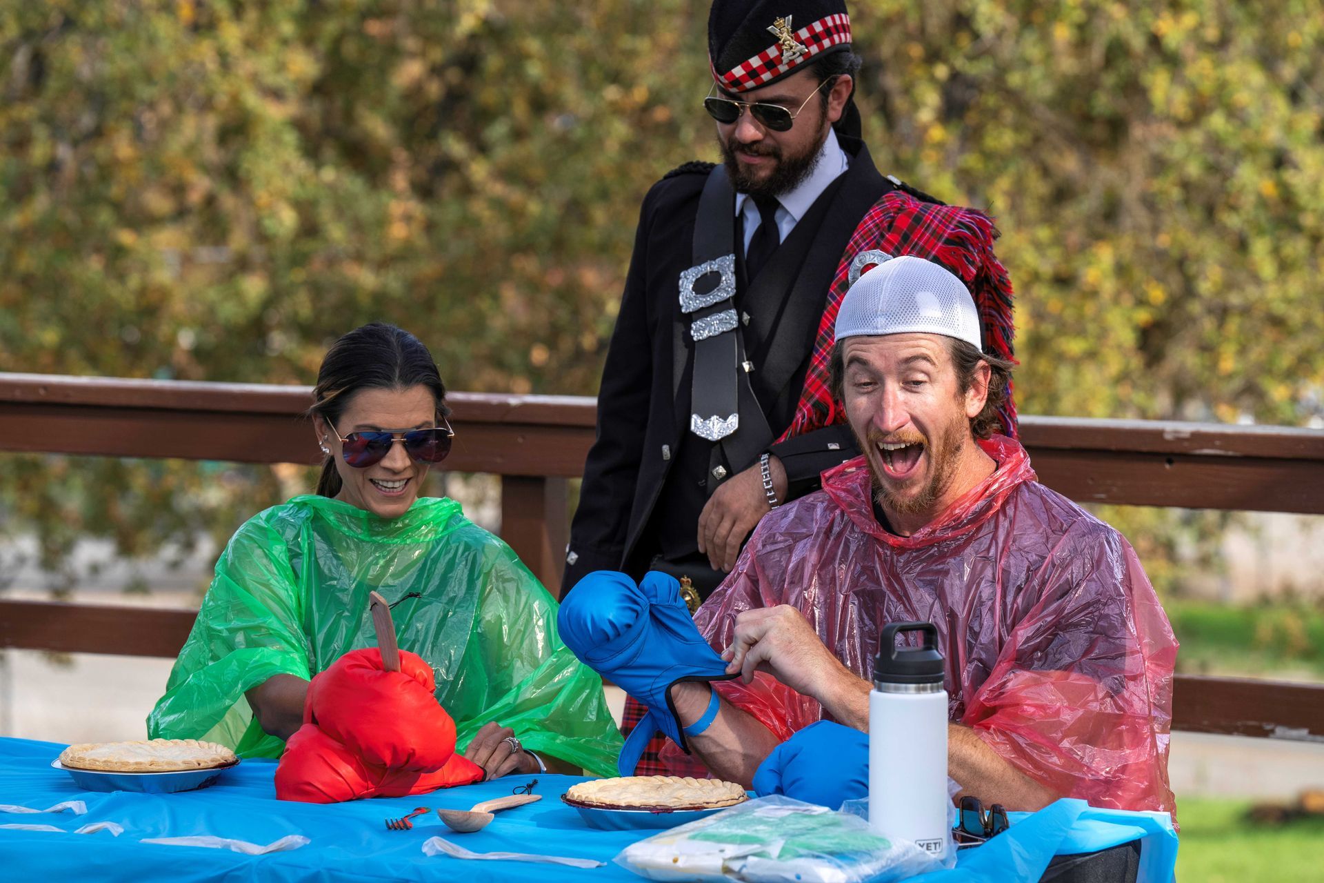 Three people laughing while covered in colorful slime at an outdoor table with bowls and a water bottle.