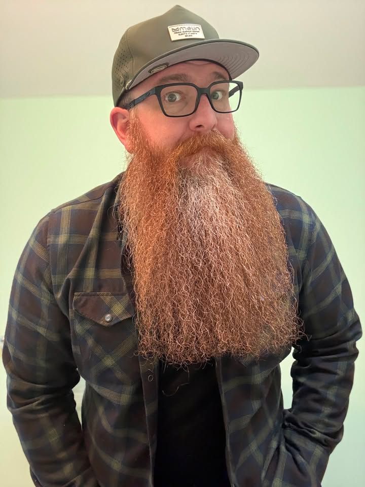 Man with glasses and a cap, wearing a plaid shirt and long beard, standing indoors against a pale wall