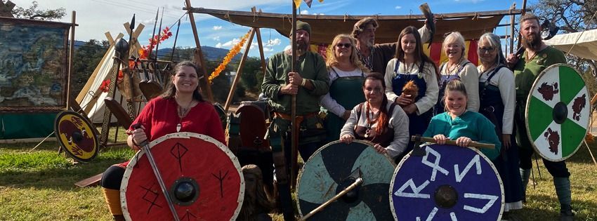 A group of people in Viking attire pose with shields. Outdoors, sunny.