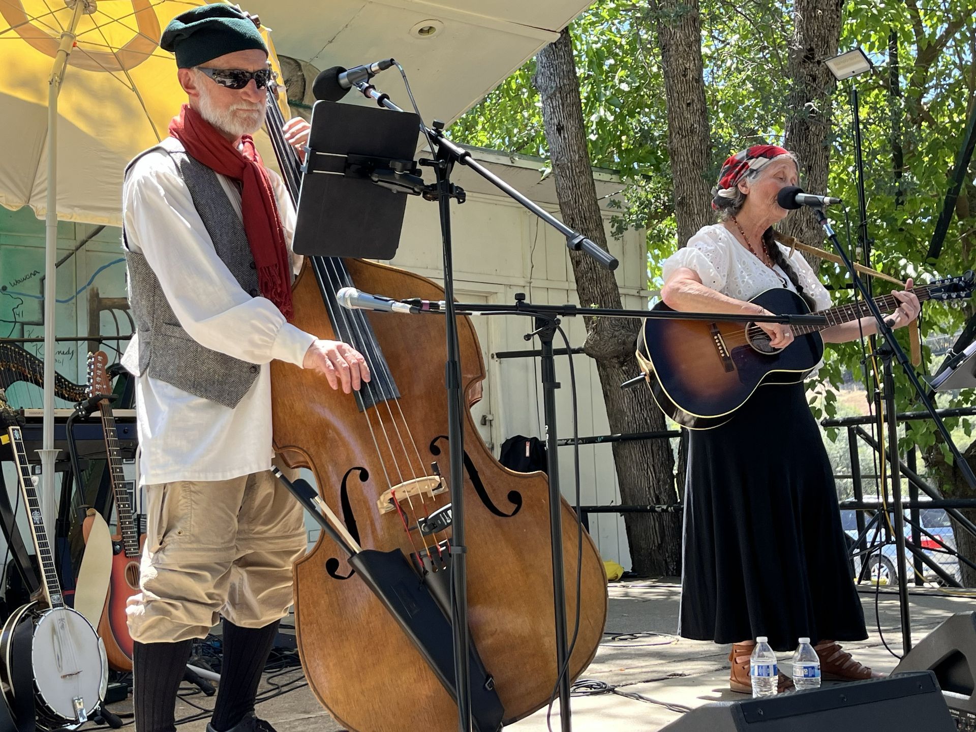Musicians performing outdoors, playing upright bass and acoustic guitar on a shaded stage.