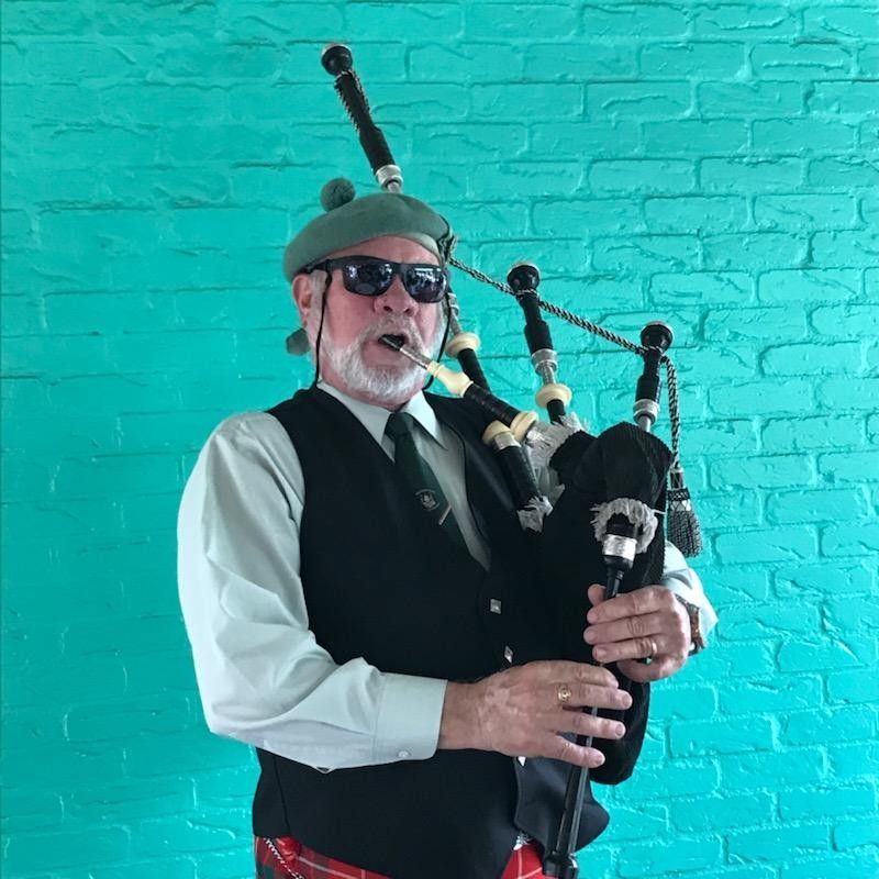 Man in Scottish attire playing bagpipes, wearing sunglasses, in front of a teal brick wall.