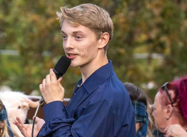 A person in a blue shirt speaks into a microphone outdoors, with a crowd in the background.