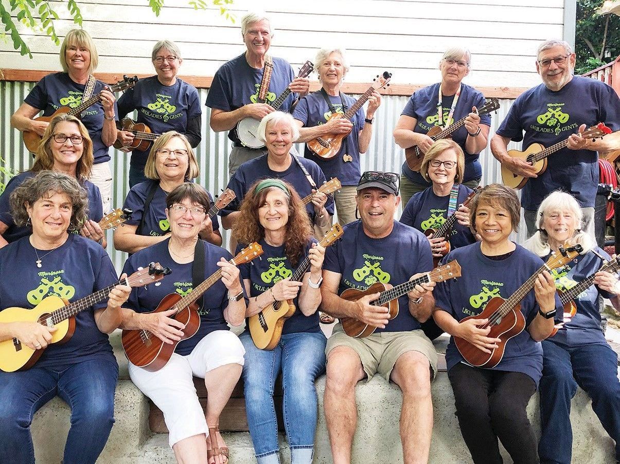 Group of people in matching blue shirts holding ukuleles outdoors, seated and standing in rows.