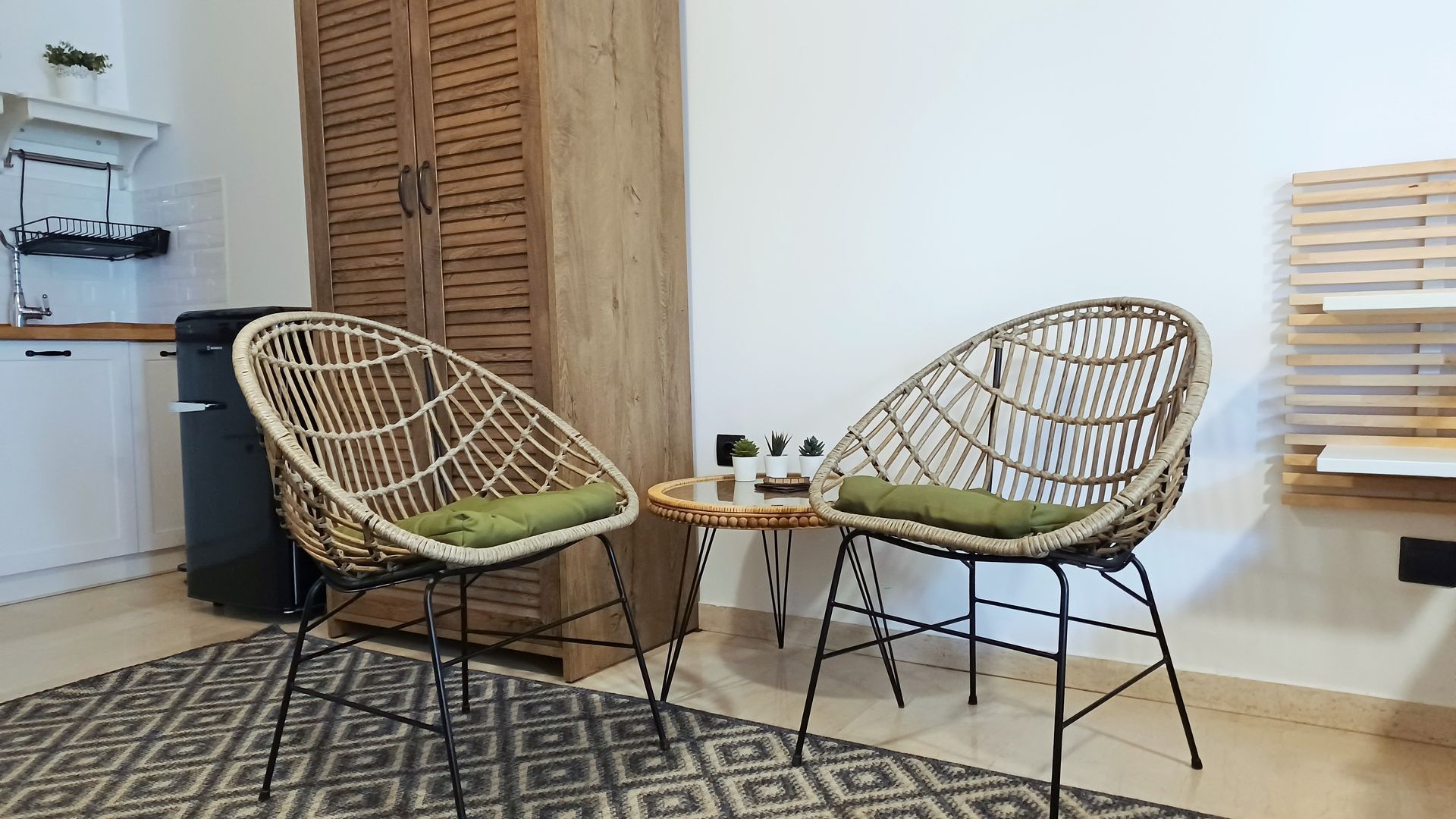 Δύο ψάθινες καρέκλες με πράσινα μαξιλάρια κάθονται πάνω σε ένα χαλί με σχέδια, κοντά σε ένα μικρό τραπέζι.