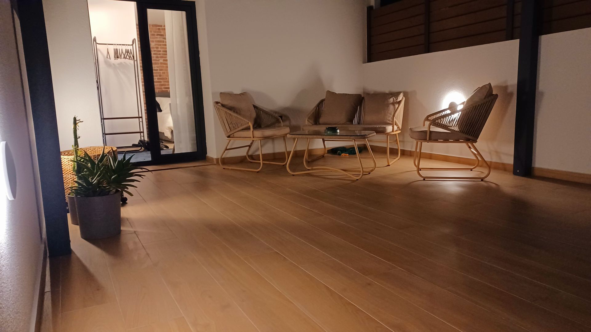 Δωμάτιο με ξύλινο δάπεδο και ψάθινα έπιπλα· πόρτα στα αριστερά, ορατά φυτά και φωτιστικό.
