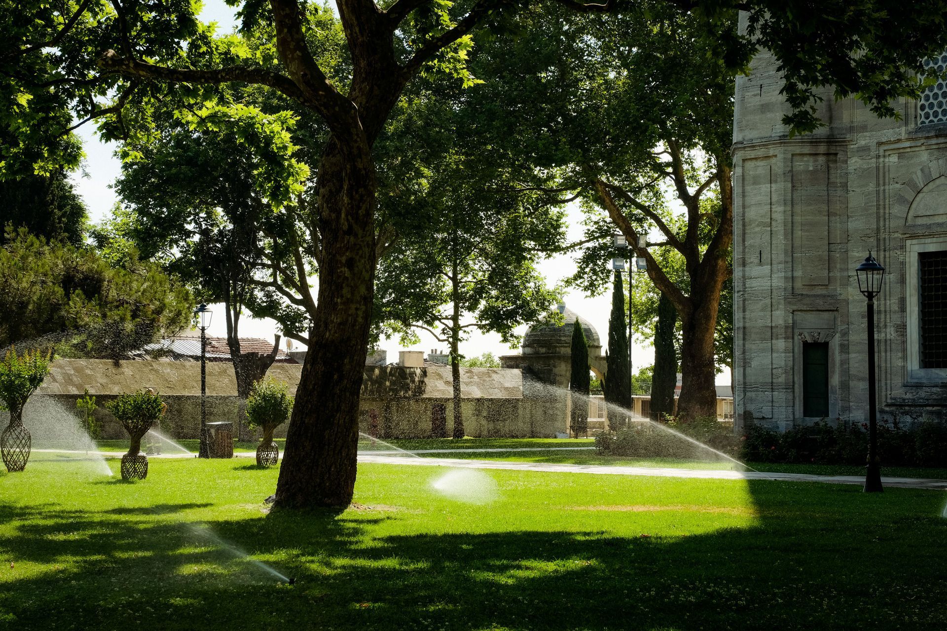 Sunny park with large trees, green lawn, and fountain sprays beside a stone building
