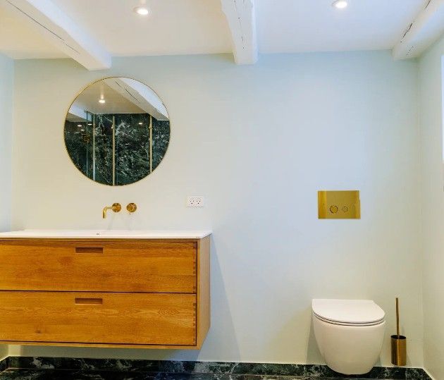 Badeværelse med rundt spejl, svævende træhåndvask, hvidt toilet og guldfarvede armaturer. Lyseblå vægge.