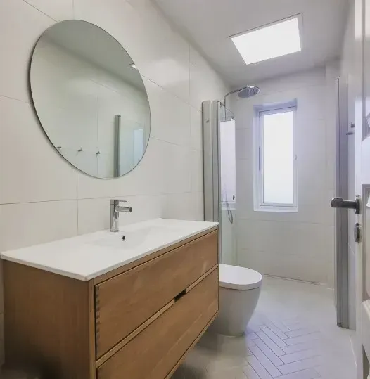 Badeværelse med træbordplade, rundt spejl og hvidt toilet. Bruseren har glasvæg.