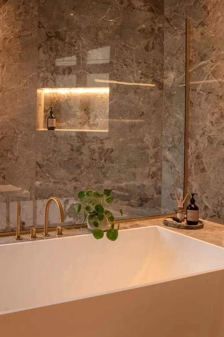 Badeværelse med marmorvægge, hvidt badekar, gylden vandhane, indbygget hylde med toiletartikler og en lille plante.