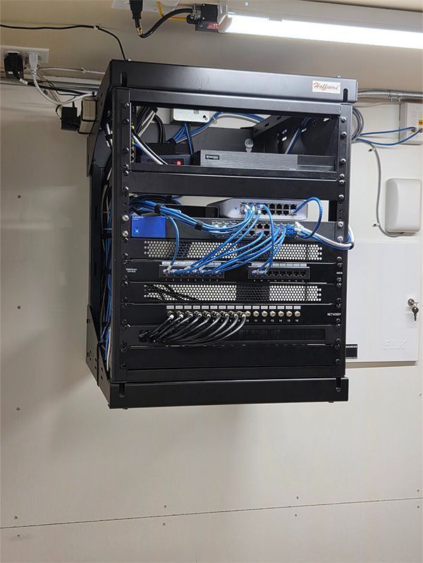 A black network rack mounted on a wall containing various network devices and blue cables.