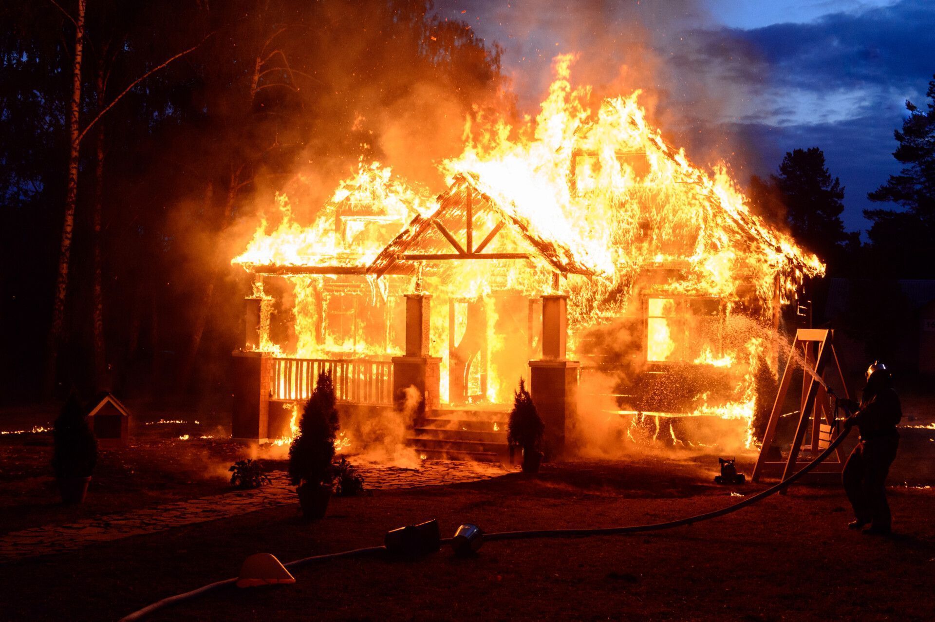 House engulfed in flames; firefighters battling fire with water hoses at night.
