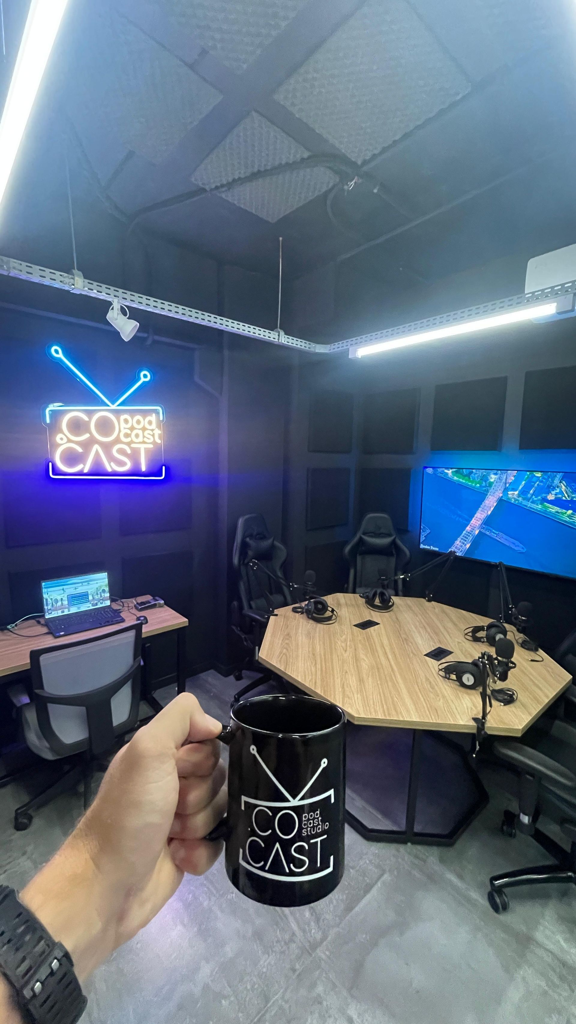 Uma pessoa está segurando uma caneca de café em uma sala com uma placa de neon no estúdio de podcast do Sandbox Coworking