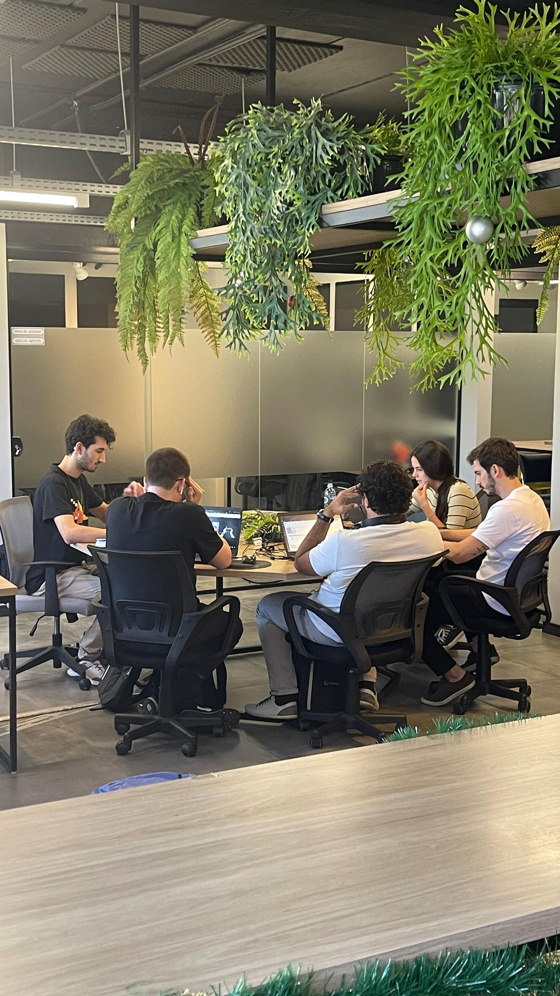 Um grupo de pessoas está sentado ao redor de uma mesa em um escritório no Sandbox Coworking