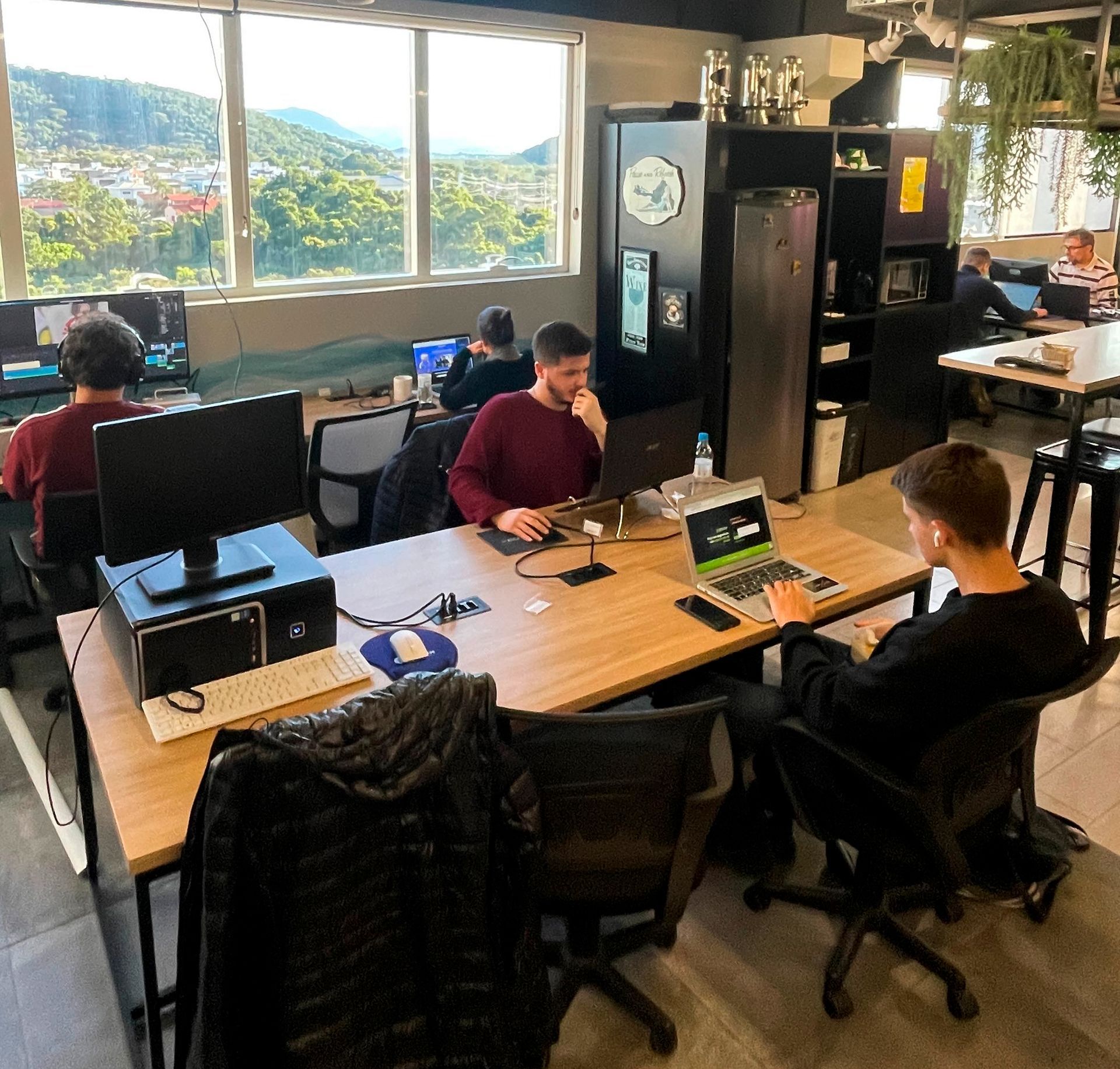 Escritório compartilhado com pessoas trabalhando em mesas, computadores e uma grande janela com vista para as colinas.