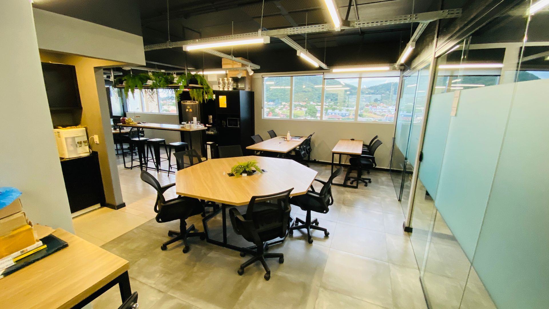 Sala de estar moderna com mesas, cadeiras pretas, paredes de vidro e grandes janelas com vista para a vegetação.