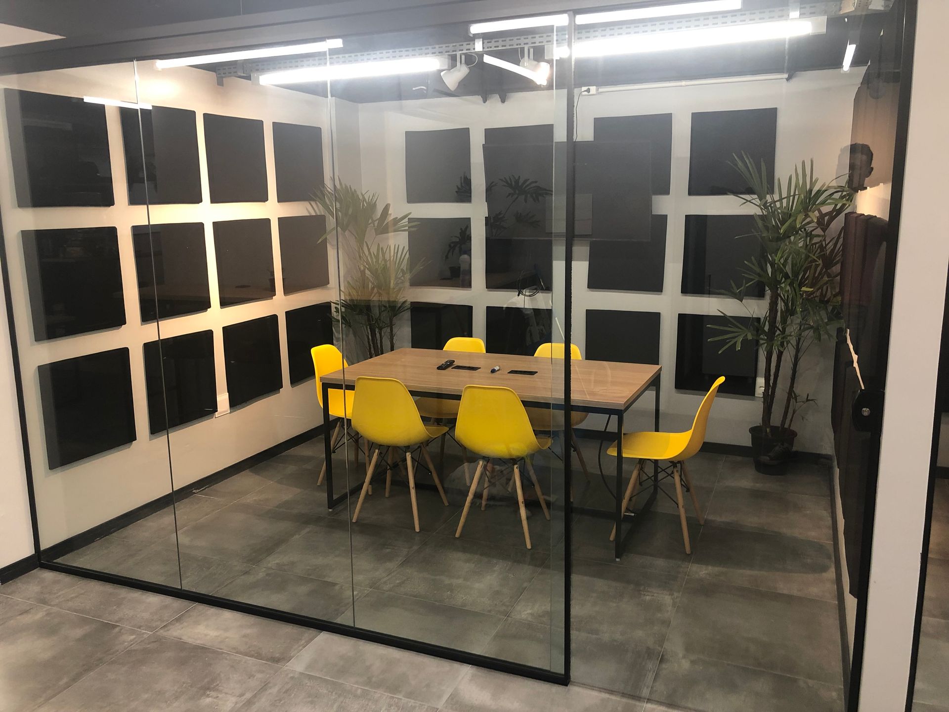 Sala de reuniões moderna com cadeiras amarelas, mesa de madeira, divisória de vidro e plantas altas.