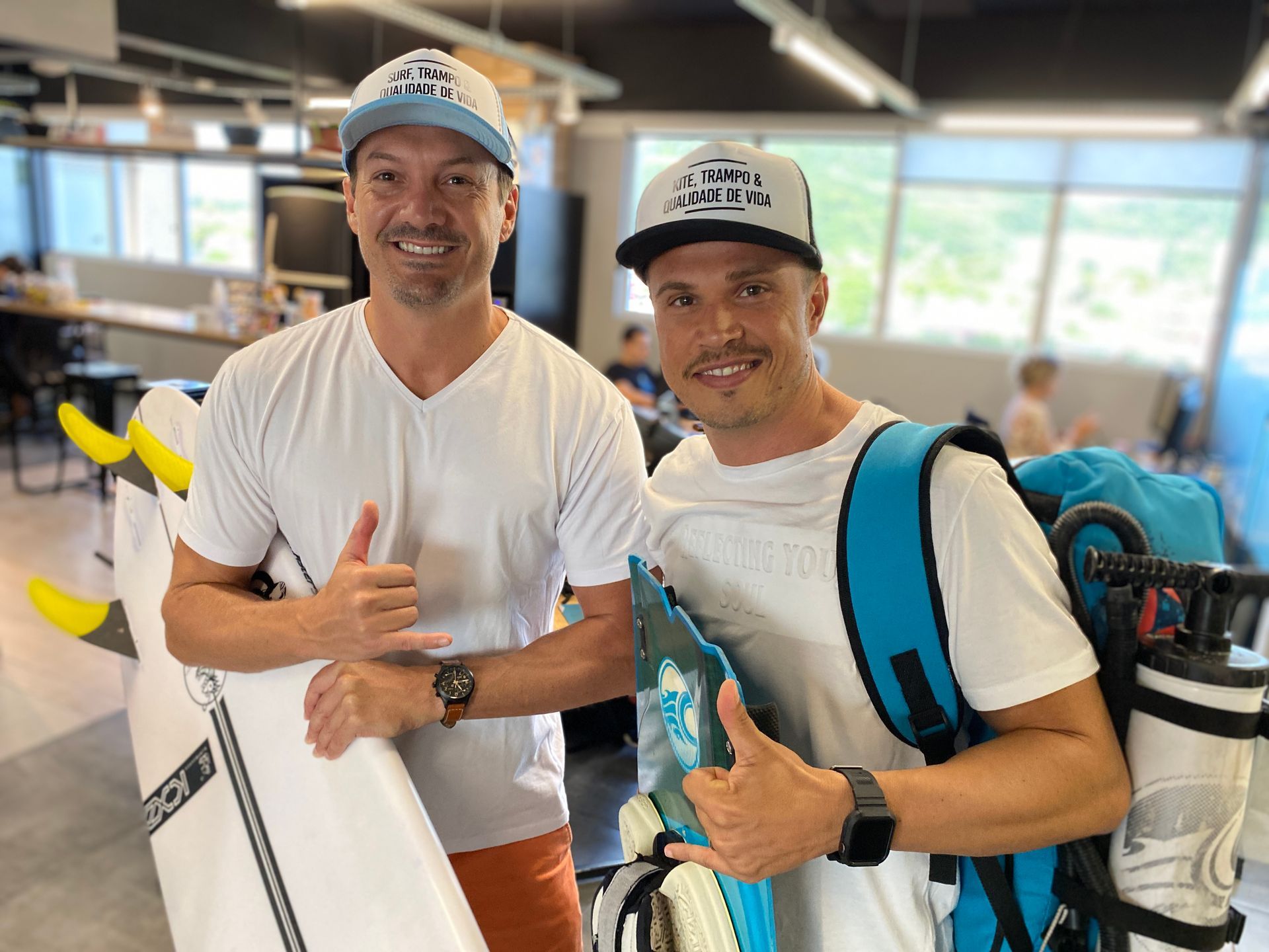 Dois homens sorrindo e fazendo sinal de positivo em um aeroporto, um deles segurando uma prancha de surfe, ambos com mochilas nos ombros.