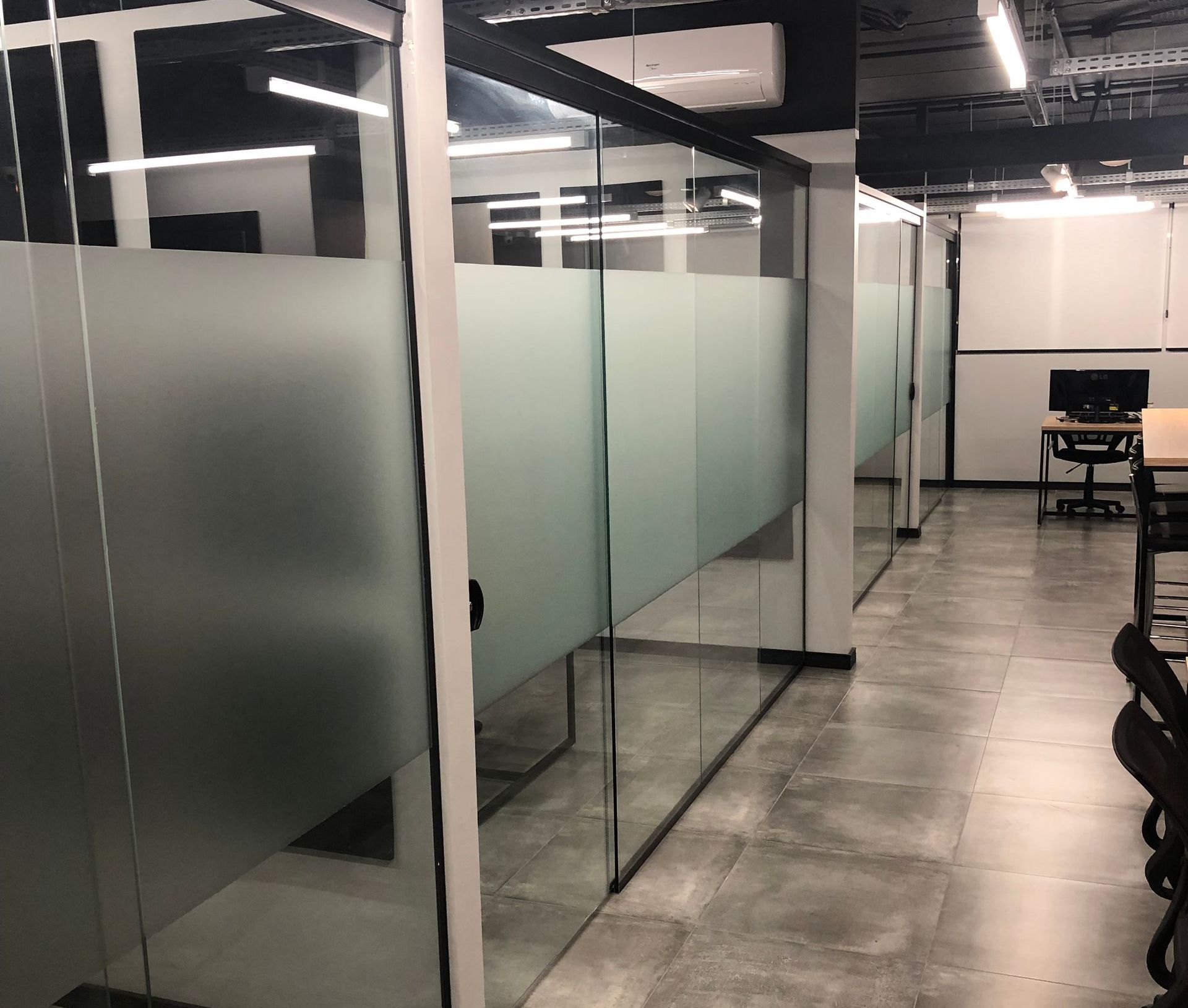 Corredor de escritório moderno e vazio, com divisórias de vidro fosco e mesas ao longo do lado direito.
