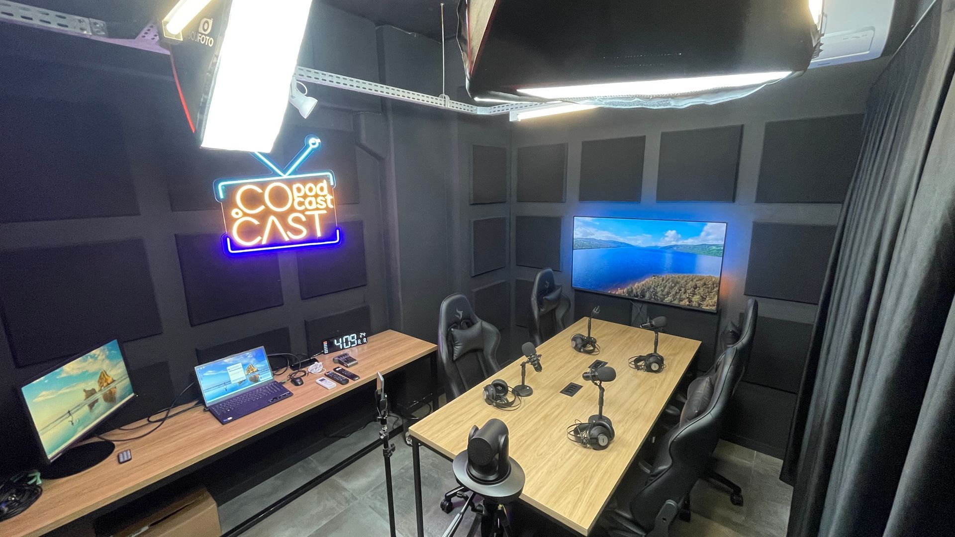 Pequeno estúdio de podcast com uma longa mesa de madeira, microfones, monitores e uma placa de néon com os dizeres “COBAS CAST”.
