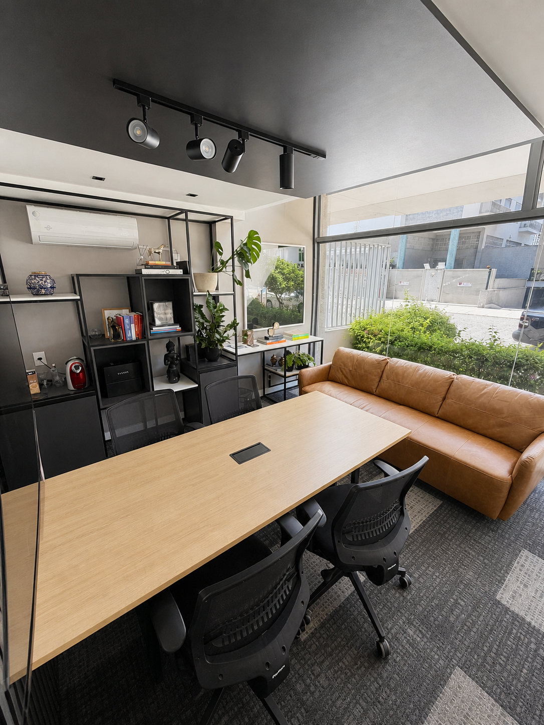 Modern meeting room with wood table, black chairs, tan sofa, shelving, and large windows