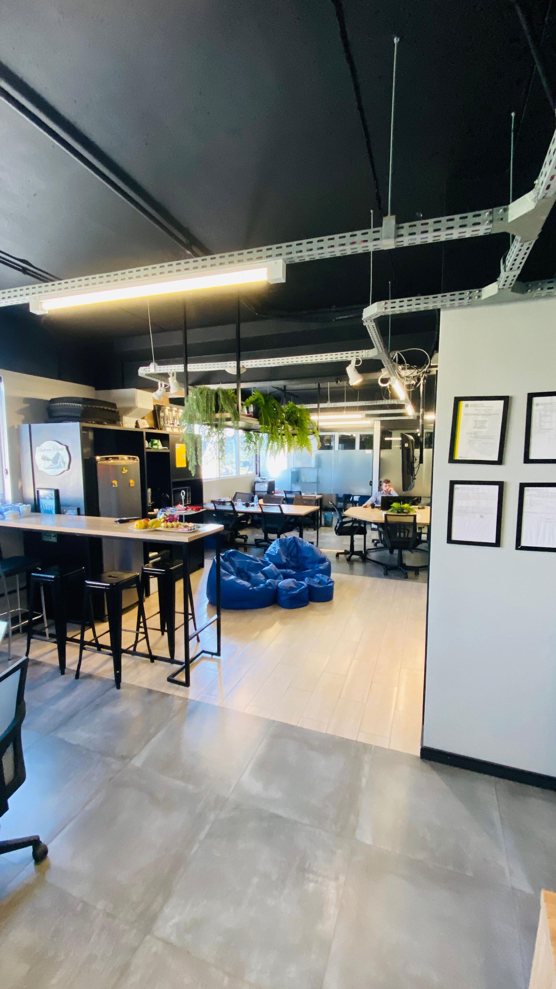 Sala de estar moderna com mesas, cadeiras azuis, plantas e obras de arte emolduradas sob iluminação aparente no teto.