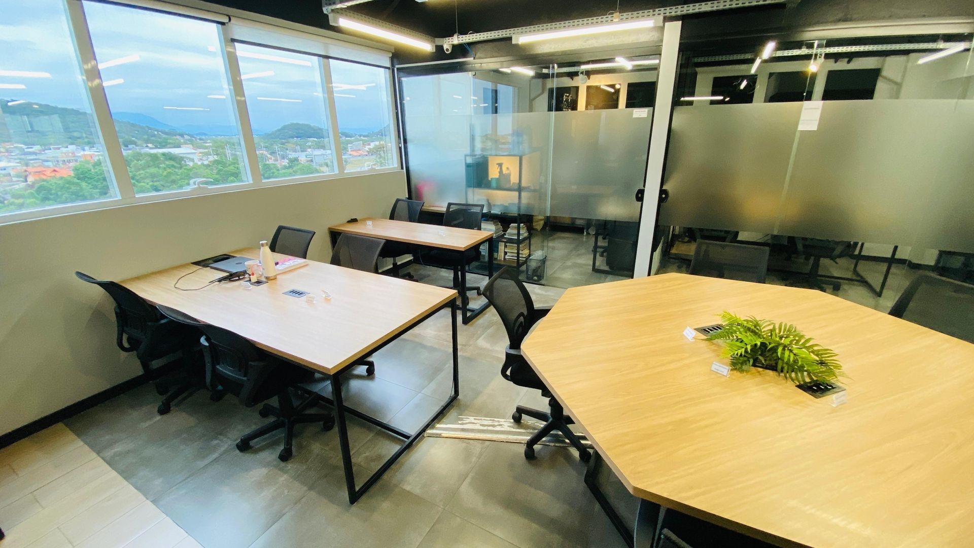 Sala de conferências moderna com mesas, cadeiras, paredes de vidro e vista para a cidade através de grandes janelas.