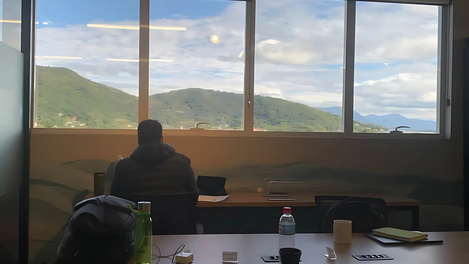 Person seated at a table, facing large window with mountain view and cloudy sky. Office setting.