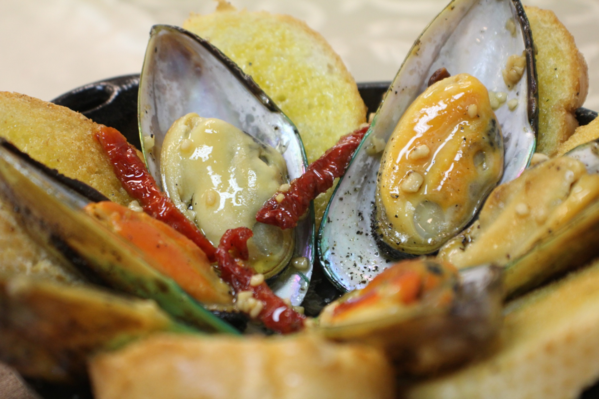A dish of New Zealand green lip mussels in a flavorful garlic wine sauce, garnished with sun-dried tomatoes. Served with crunchy garlic crostini on the side.