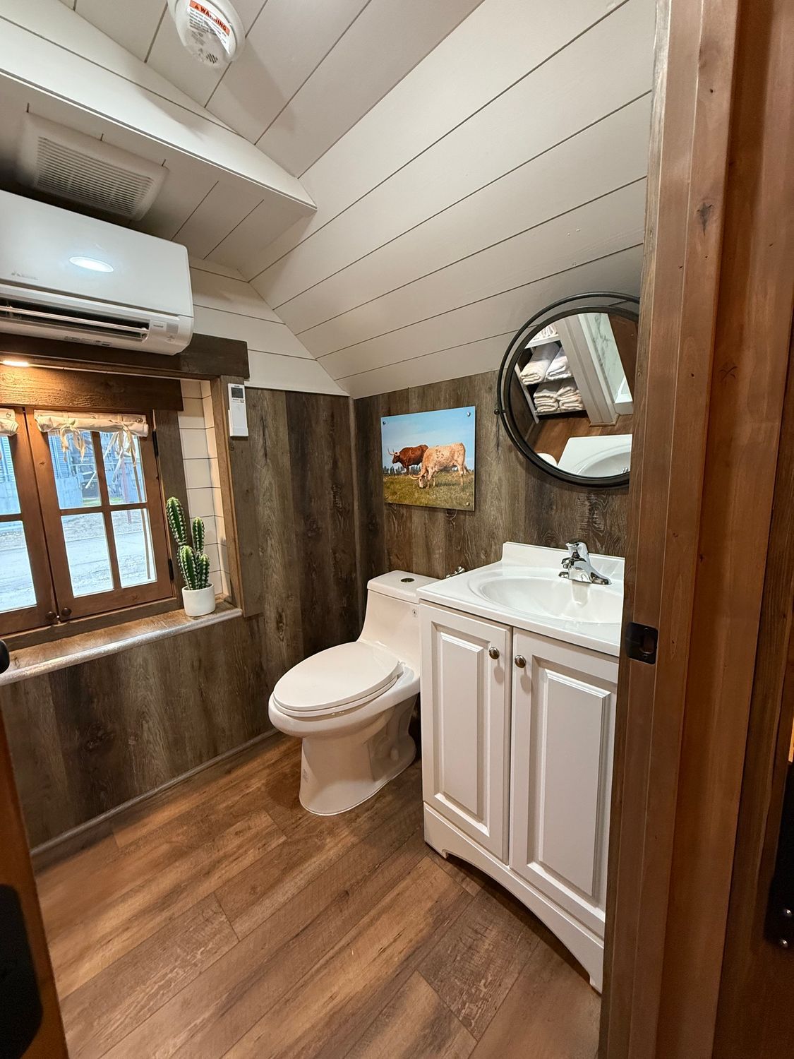 Compact rustic bathroom with white vanity, toilet, wood walls, and window air conditioner