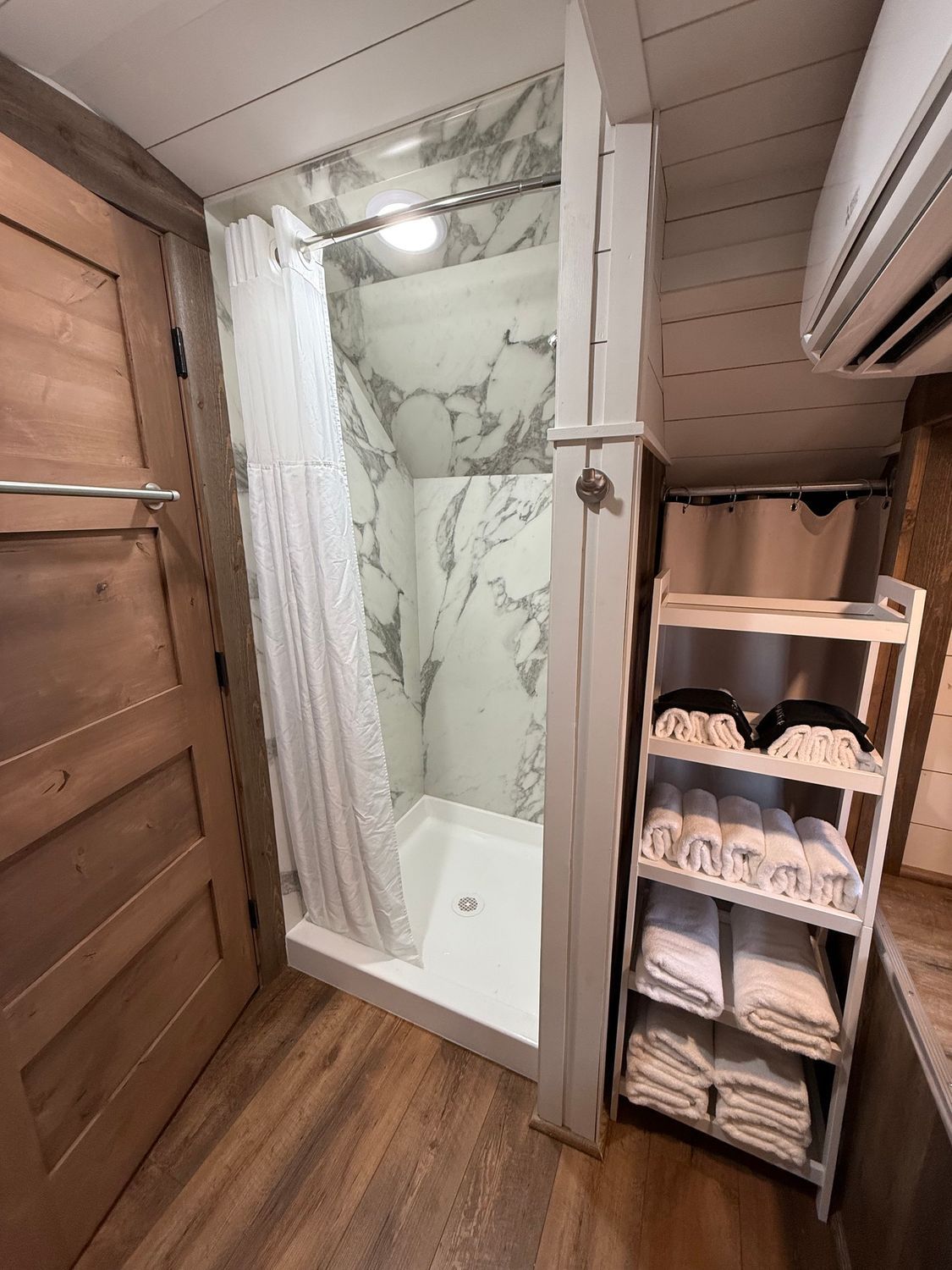 Narrow RV bathroom with shower curtain, marble-pattern shower wall, and towel shelf beside the door