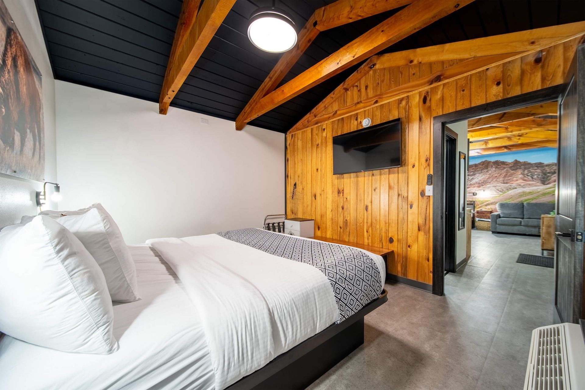 Modern bedroom with white bedding, wood-paneled wall, dark ceiling beams, and a TV opposite the bed
