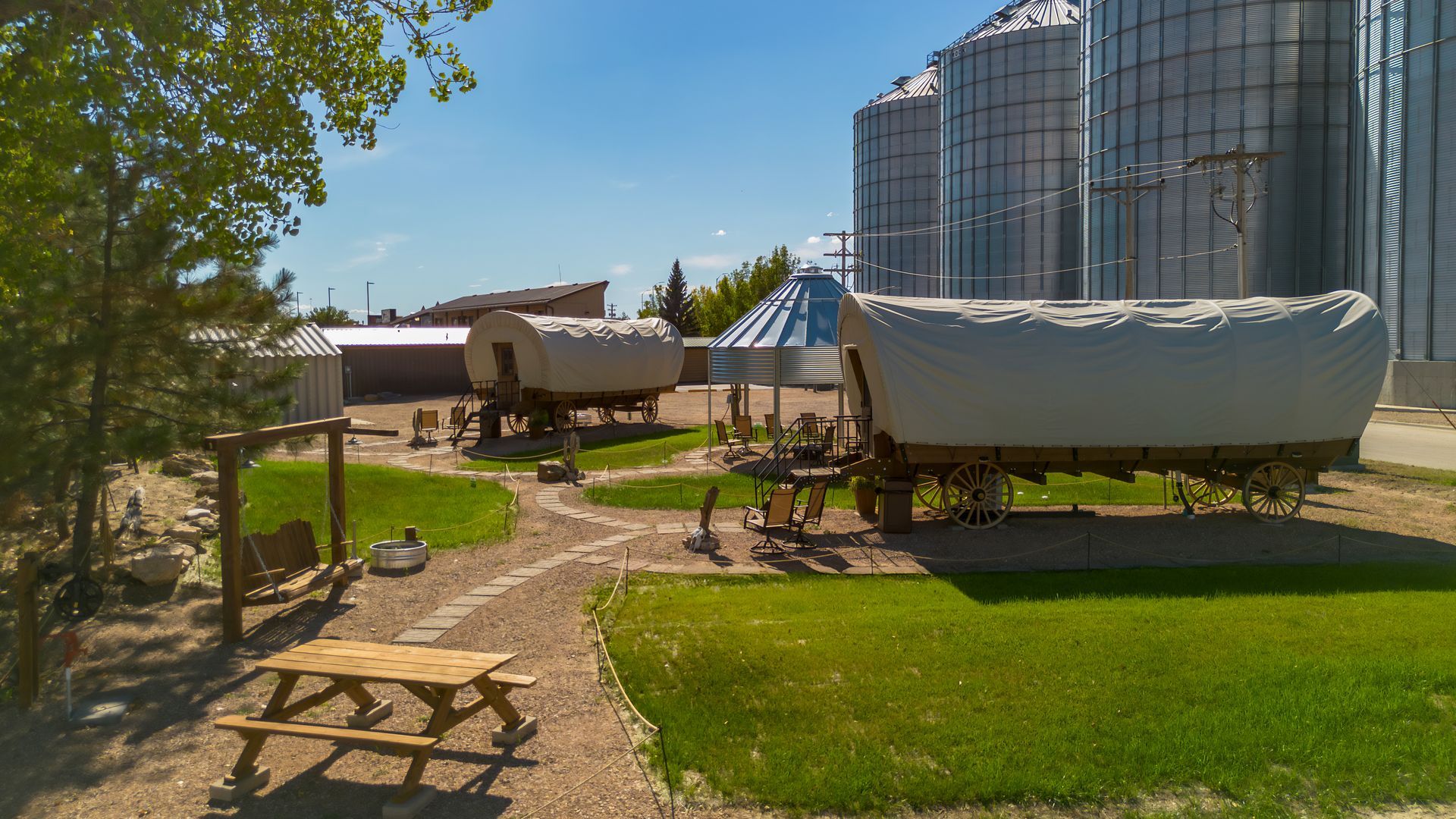 Farmyard with covered wagons, grain silos, and a shaded picnic table beside a dirt path.