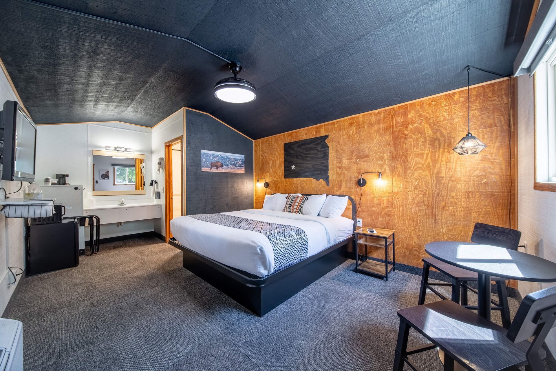 Modern hotel room with bed, desk, chairs, and warm wood-paneled walls.