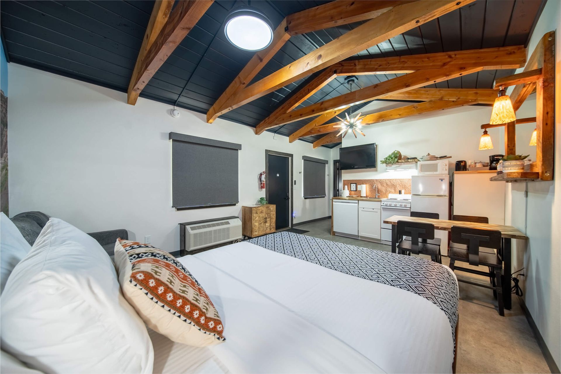 Cozy loft bedroom with exposed wood beams, bed, desk, and small kitchenette under sloped ceiling