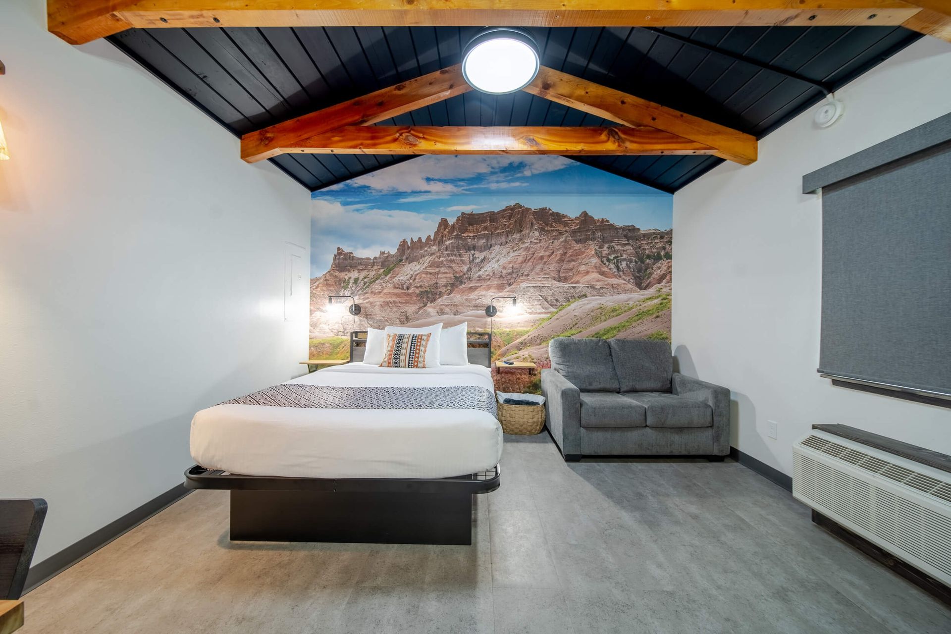 Cozy bedroom with bed, gray sofa, mountain mural, and exposed wood ceiling