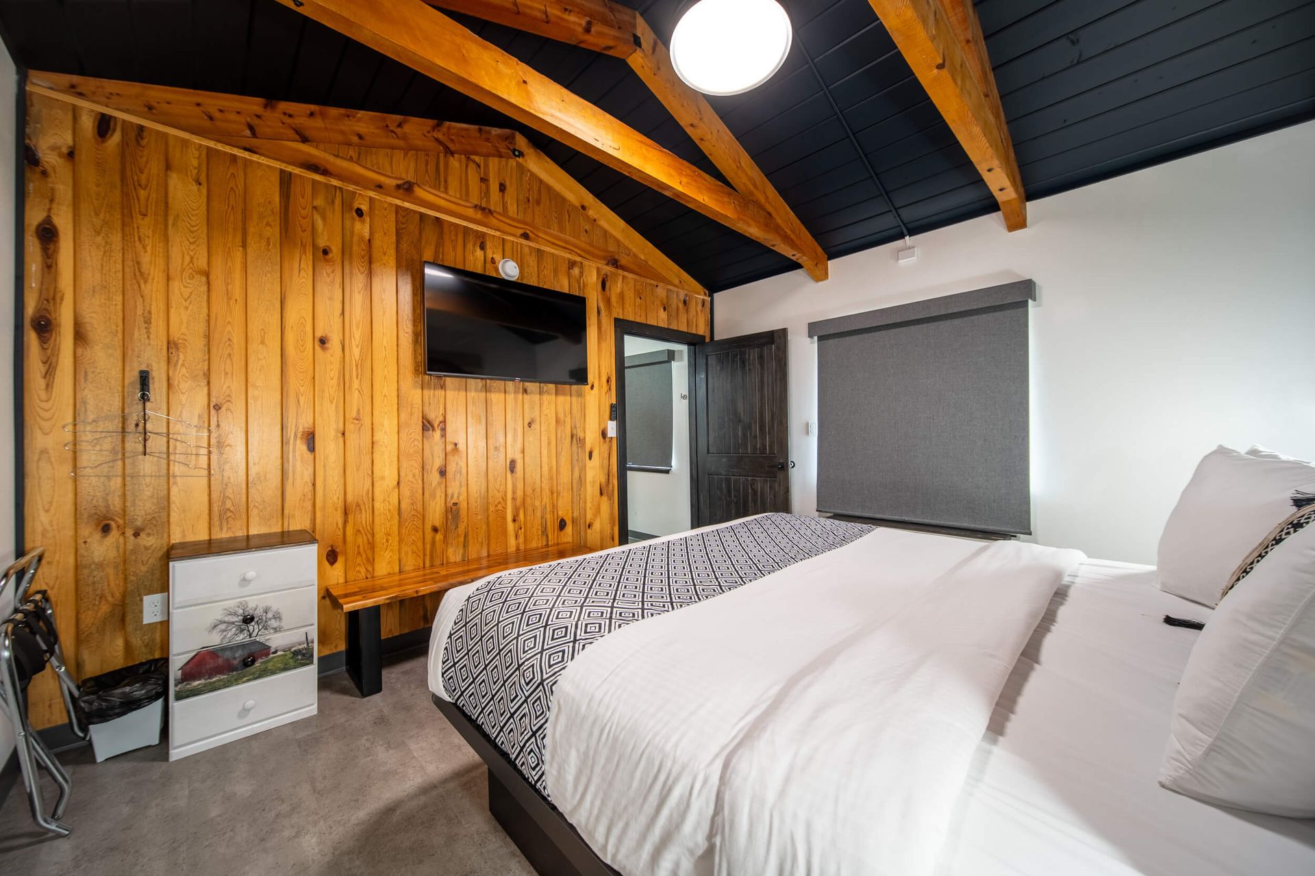 Modern bedroom with wood-paneled wall, exposed beams, a bed, and a large window with blinds.