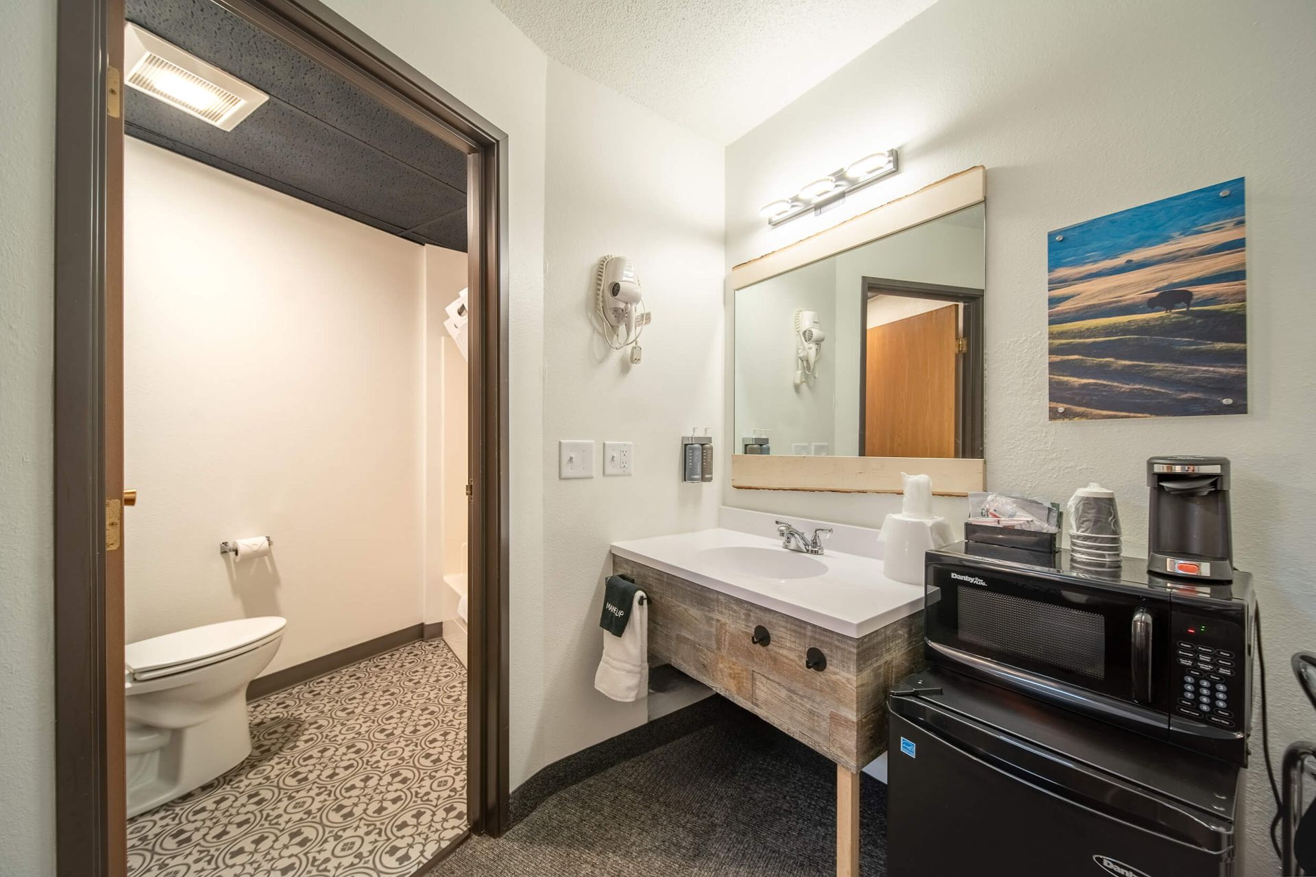 Compact restroom with beige walls, a white sink, mirror, and a toilet visible in the adjoining stall.
