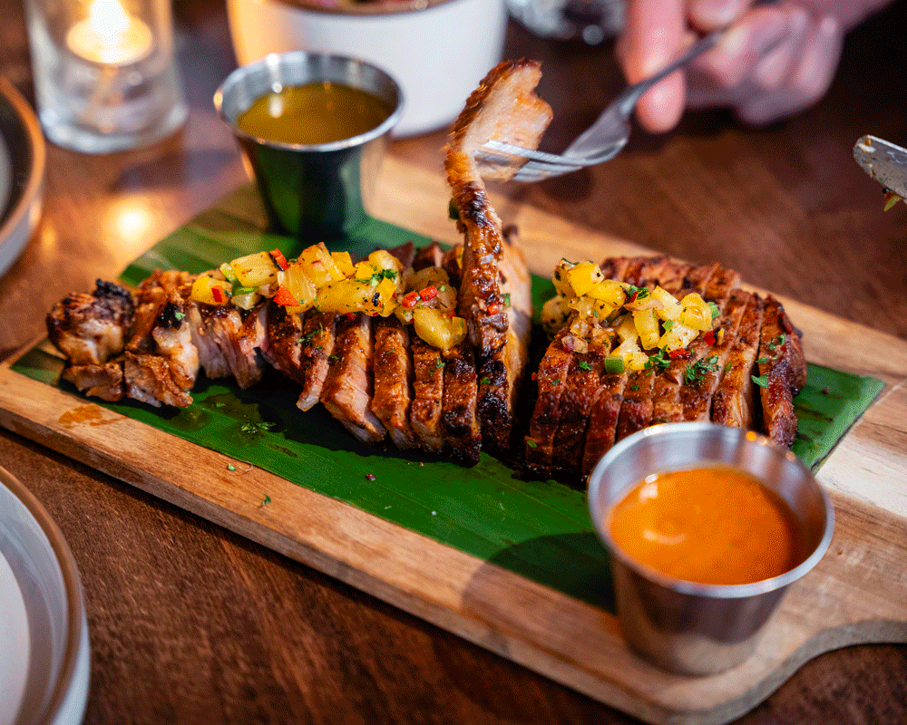 Grilled fish topped with mango salsa on a banana leaf, served with two dipping sauces on a wooden board