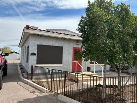 Small white house with red door and black window shutters behind a black fence, with a tree in front.