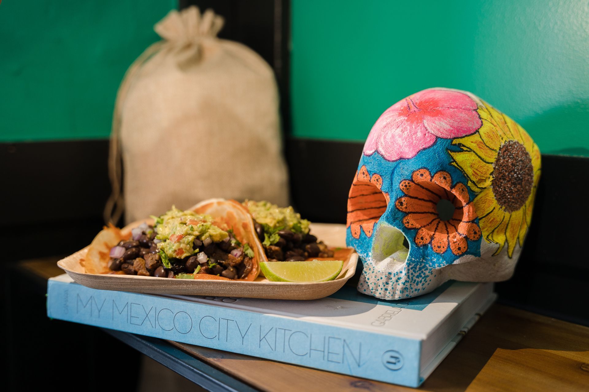Colorful floral skull beside tacos on a blue tray against a green and black background