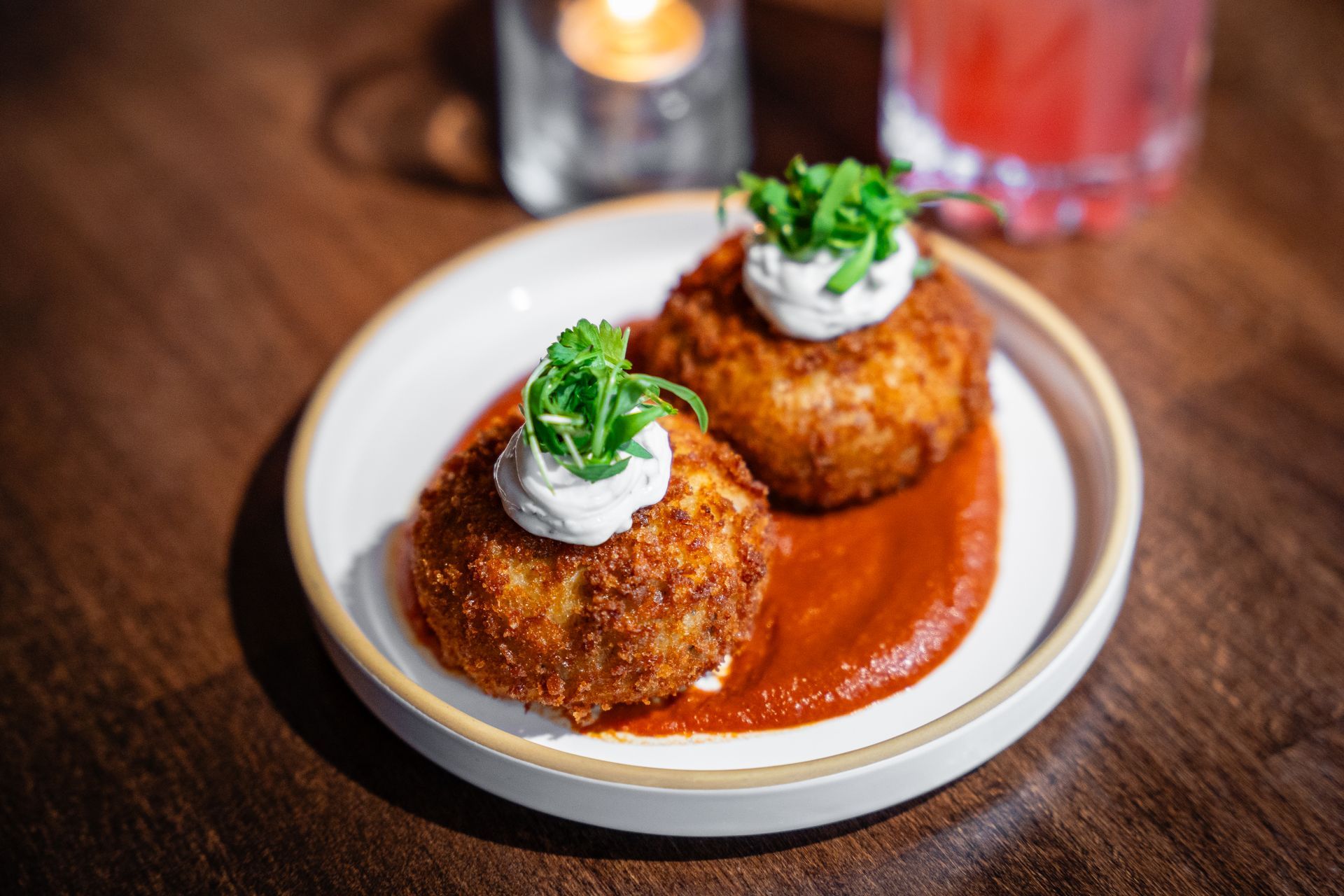 Two breaded croquettes topped with cream and herbs on a white plate with red sauce