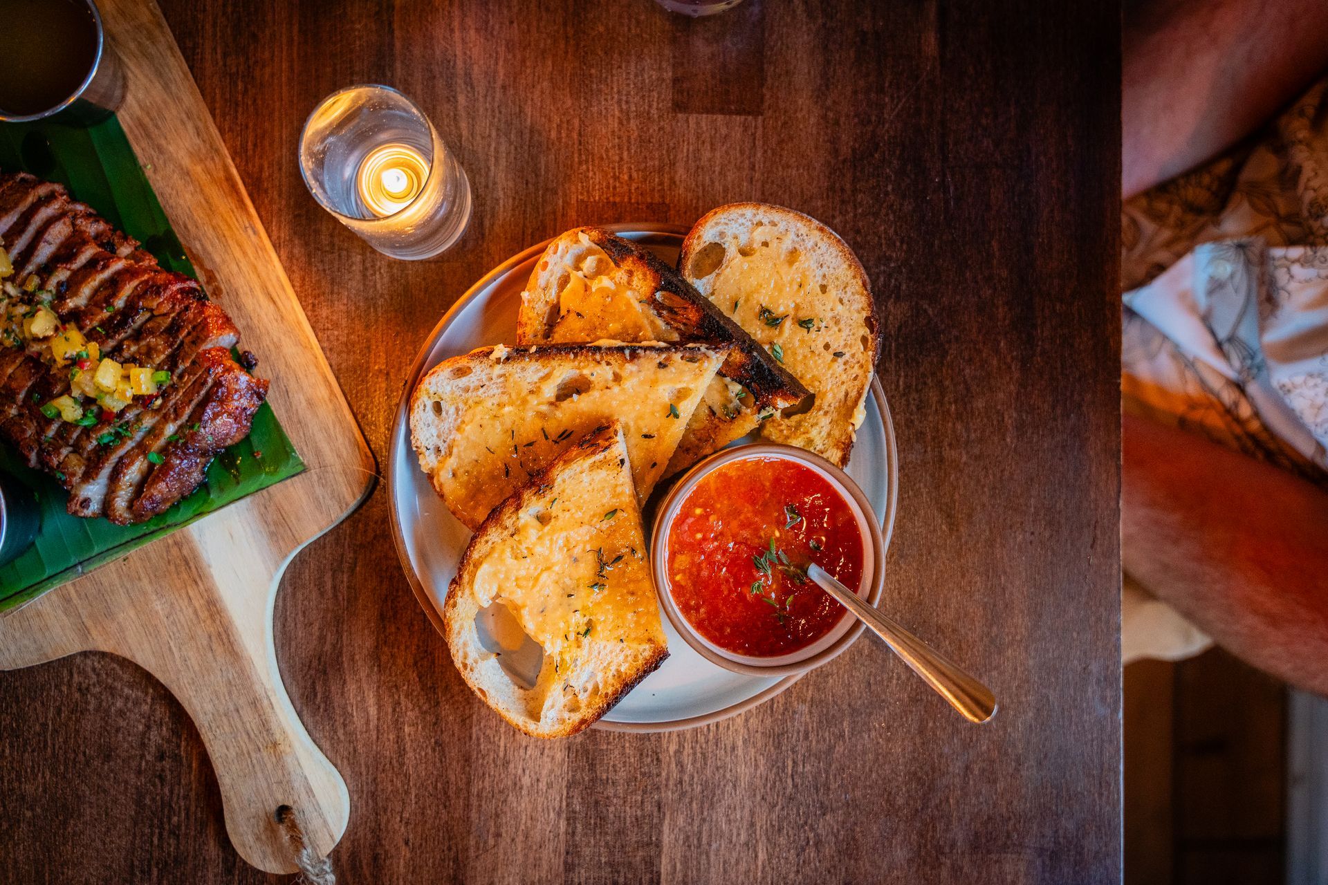 Bread and tomato soup on a wooden table beside a lit candle and grilled dish