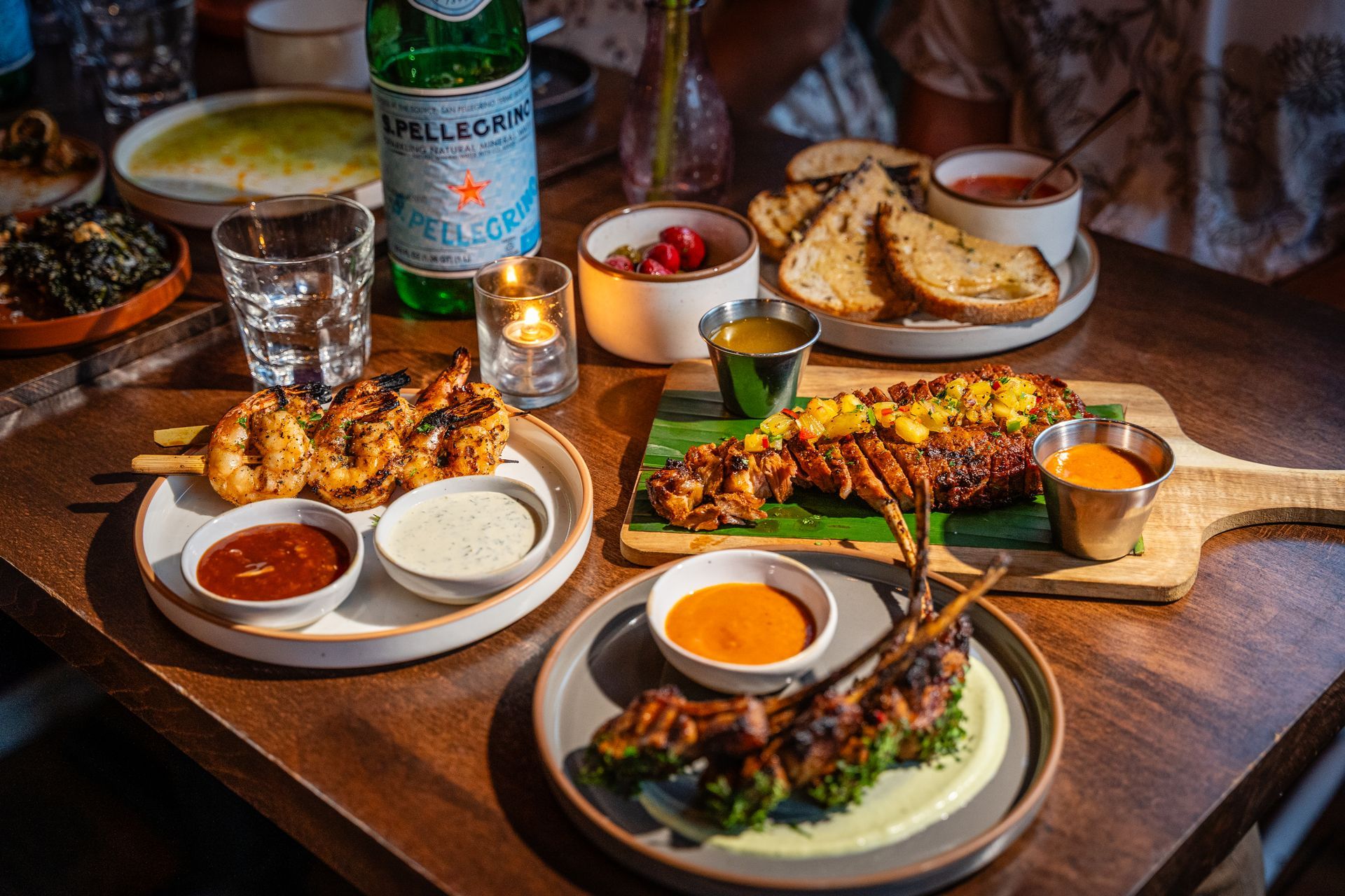 Assorted grilled dishes and dipping sauces on a wooden table with drinks and bread at a restaurant.