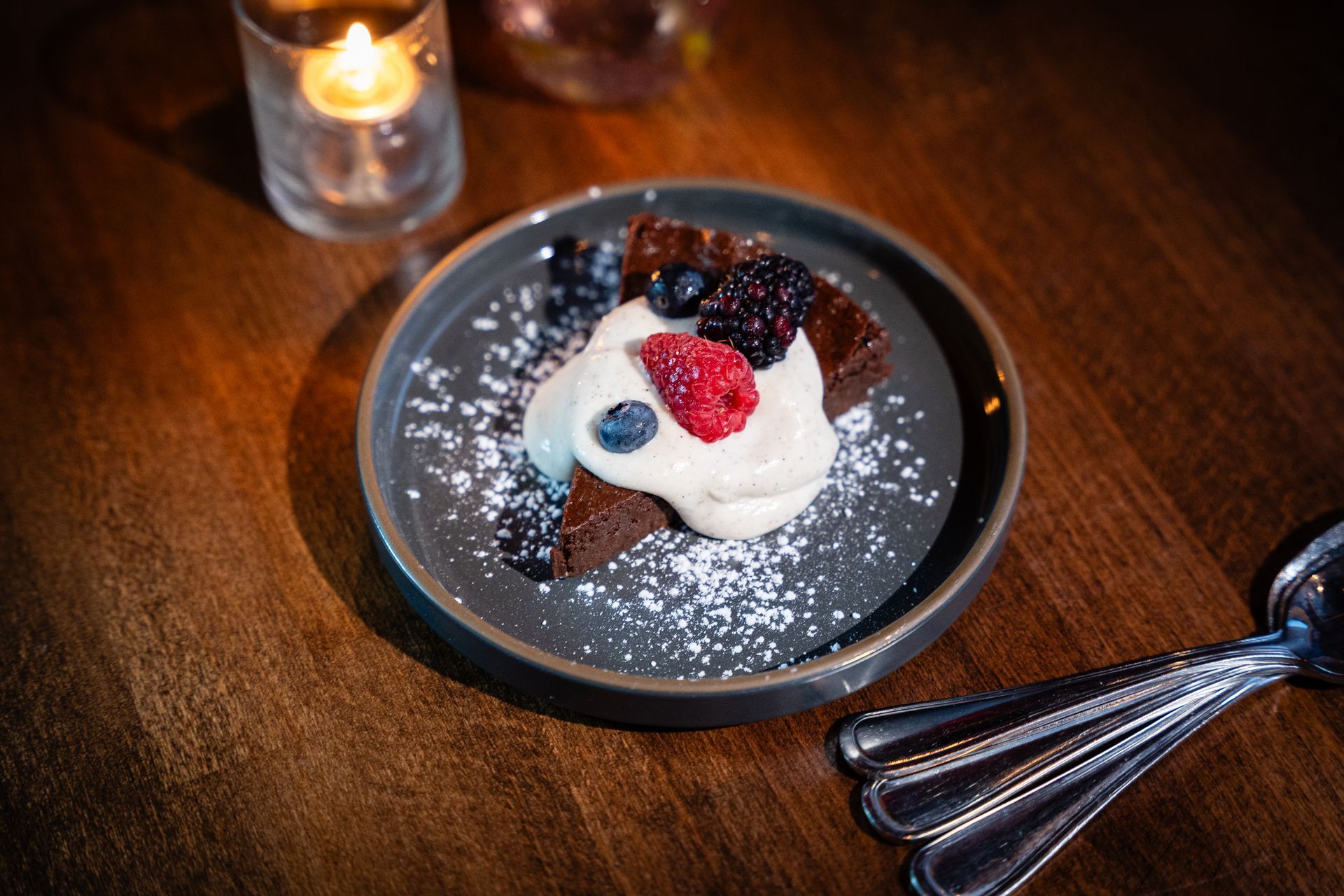Dessert on a black plate with berries and cream on a wooden table beside a candle and fork