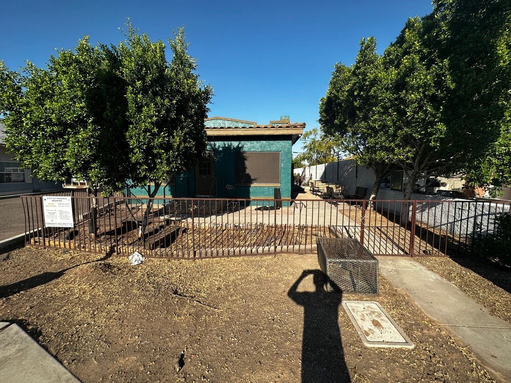 Fenced yard with two leafy trees, a small building, and a clear blue sky.