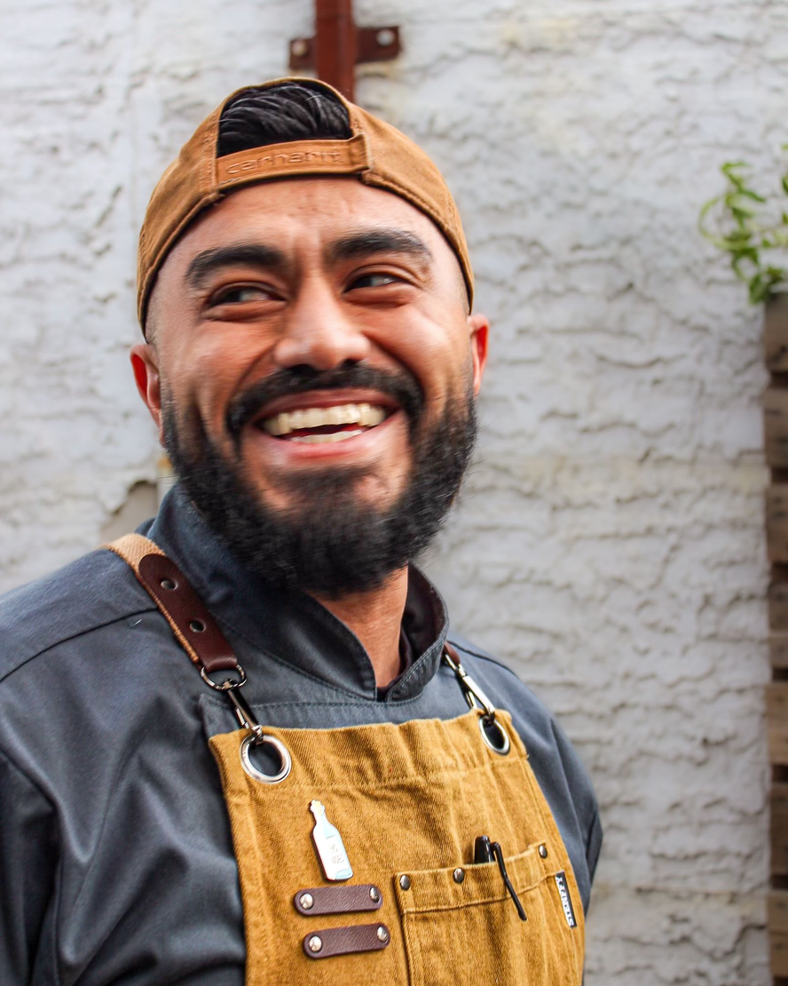 Smiling man in a tan cap and apron, standing against a light brick wall.