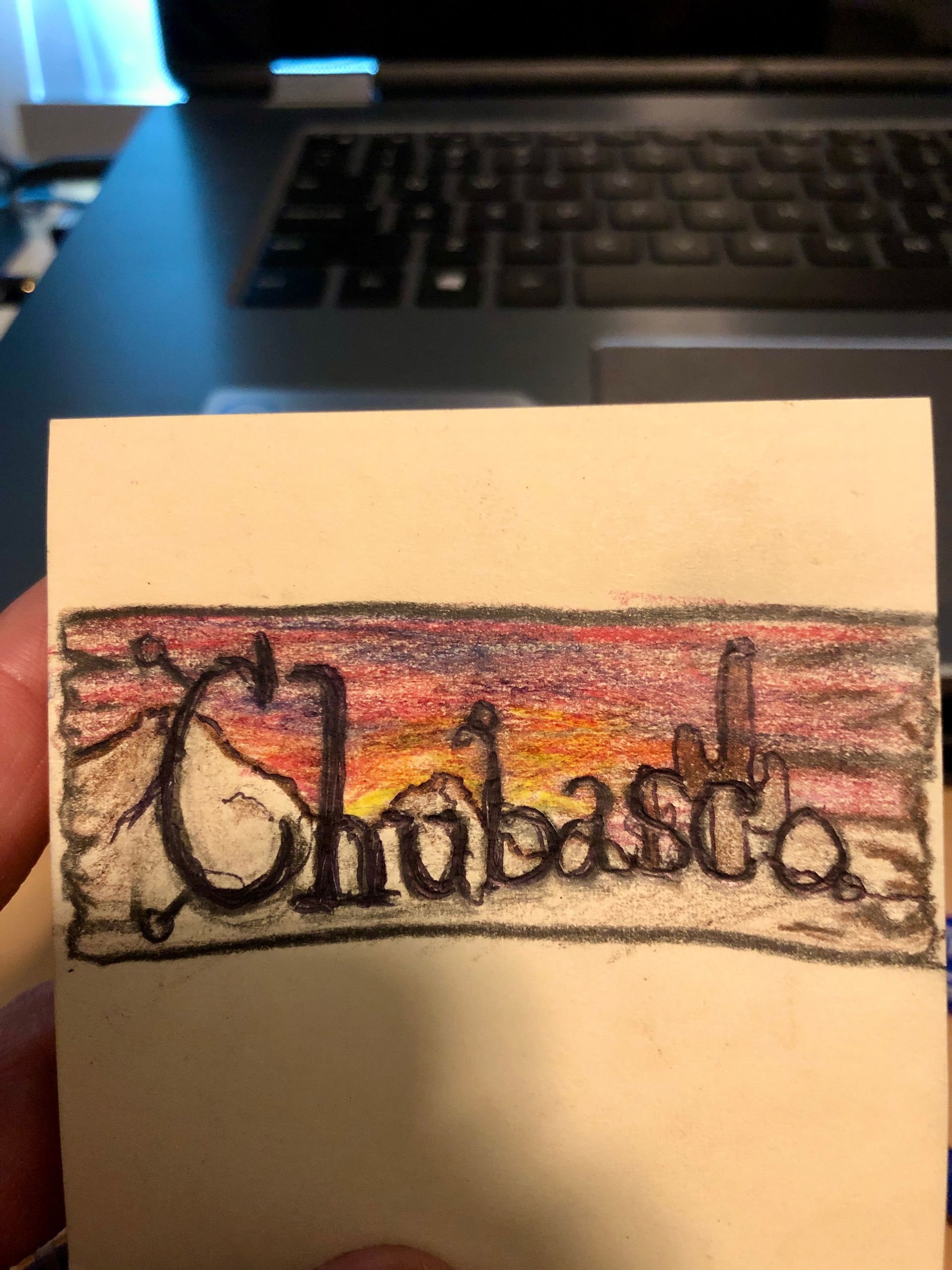 Hand-drawn “Chubaska” logo on a small paper card, held in front of a laptop keyboard